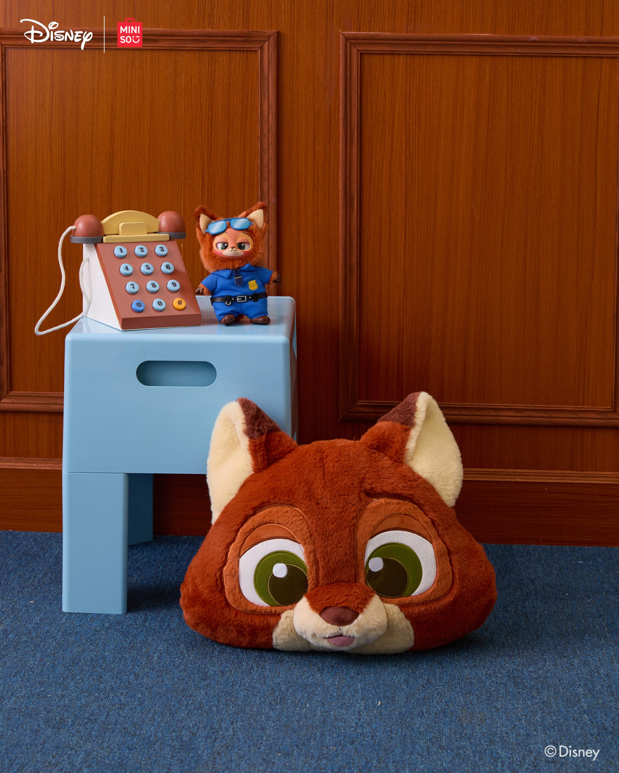A plush fox face pillow rests on blue carpet beside Zootopia 2 merchandise on a small blue table, evoking Disney park vibes.