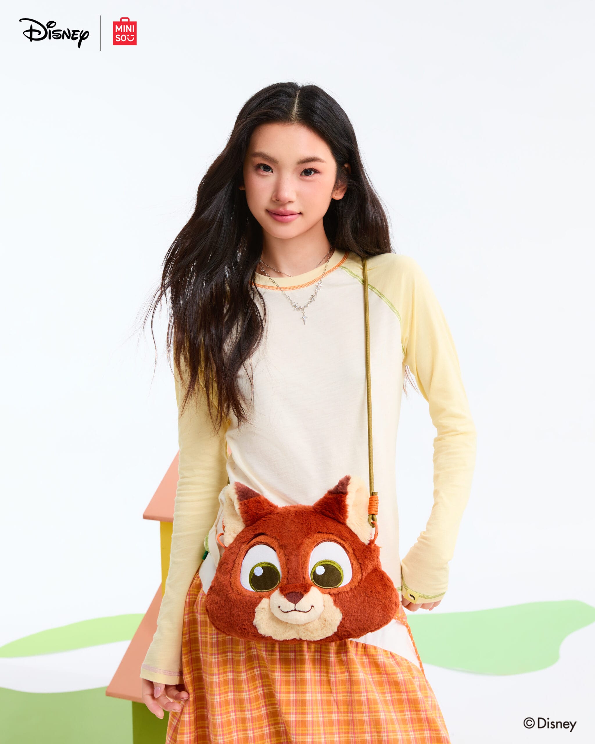A young woman smiles at a Disney park, wearing a cream shirt, plaid skirt, and plush red panda Disney bag—castle not in view.