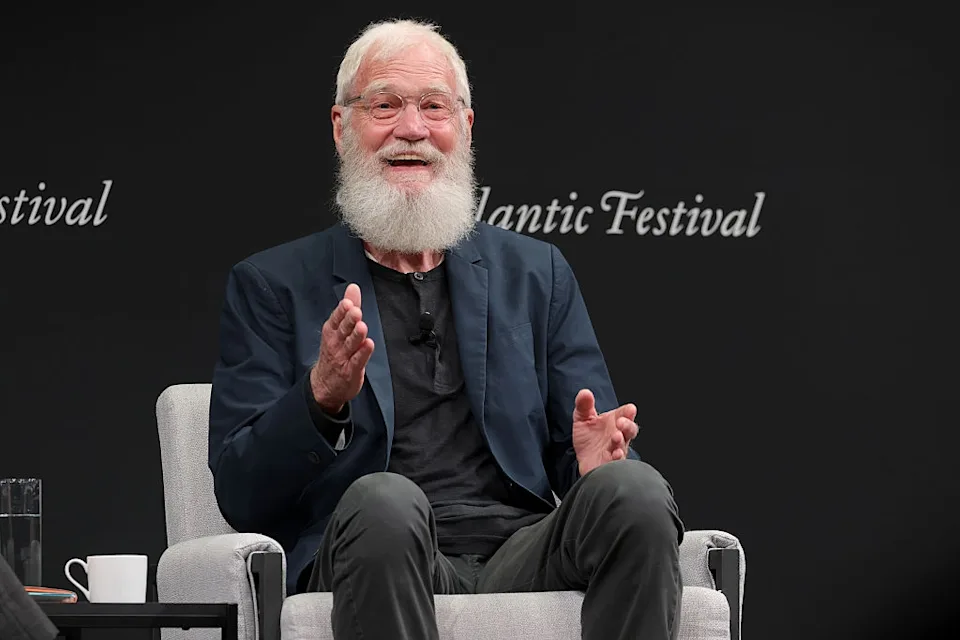 Person with a white beard, wearing a suit, sits on stage speaking, at an event with 