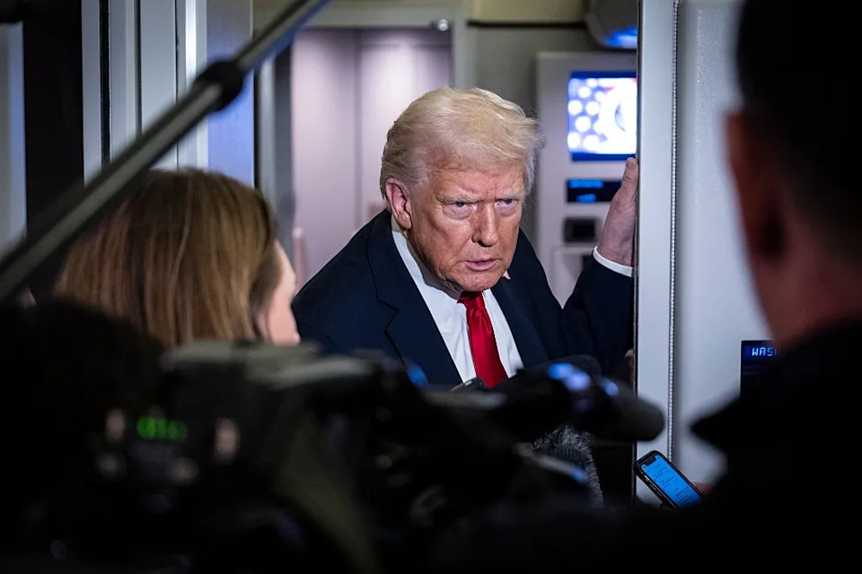 A person with styled hair and a red tie stands in a crowded press space, facing camera lenses and reporters