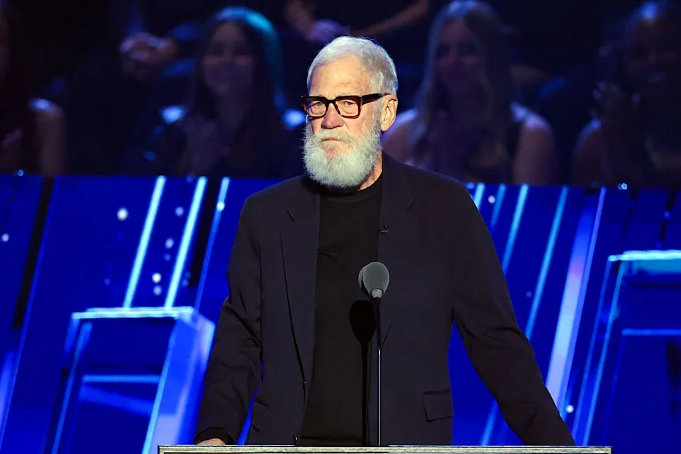 Man with a white beard and glasses speaking at an event, wearing a dark suit with a black shirt, in front of a futuristic backdrop