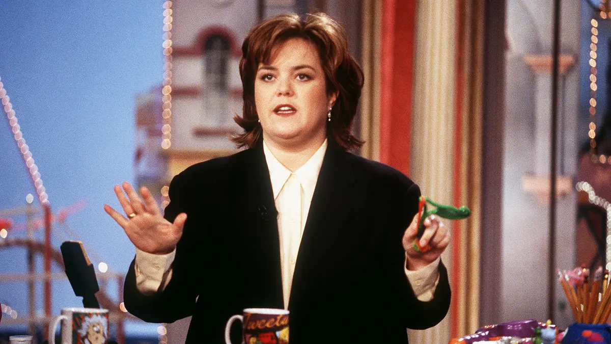 Rosie O'Donnell in a black suit puts her hands up as she hosts her talk show
