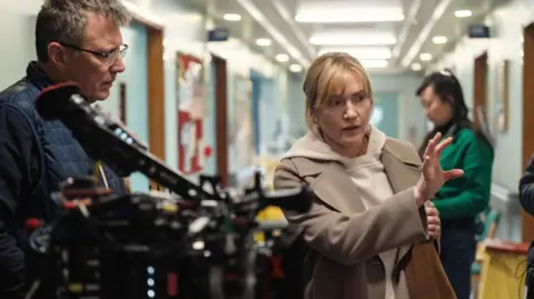 Netflix Winslet (right) directing Goodbye June, she is wearing a taupe coat and is pictured alongside a camera operator