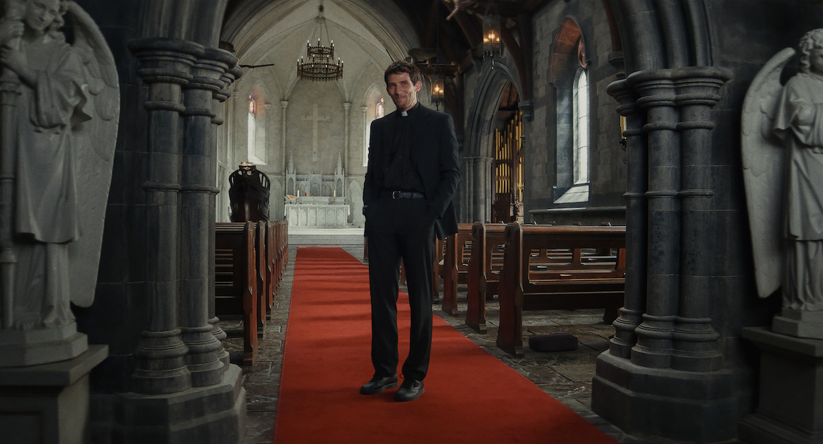 A priest stands on a red carpet in the middle aisle of a dimly lit stone church, flanked by statues, with pews on either side and an altar in the background, creating a solemn and contemplative mood.