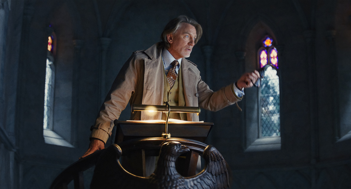 An older man in formal attire stands at a lectern shaped like an eagle in a dimly lit, gothic-style room with stained glass windows, creating a serious and dramatic atmosphere.