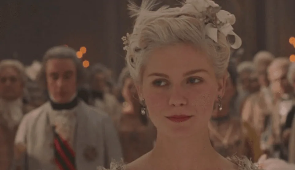 A woman in elegant historical attire at a formal gathering, with guests in the background. | IMDB