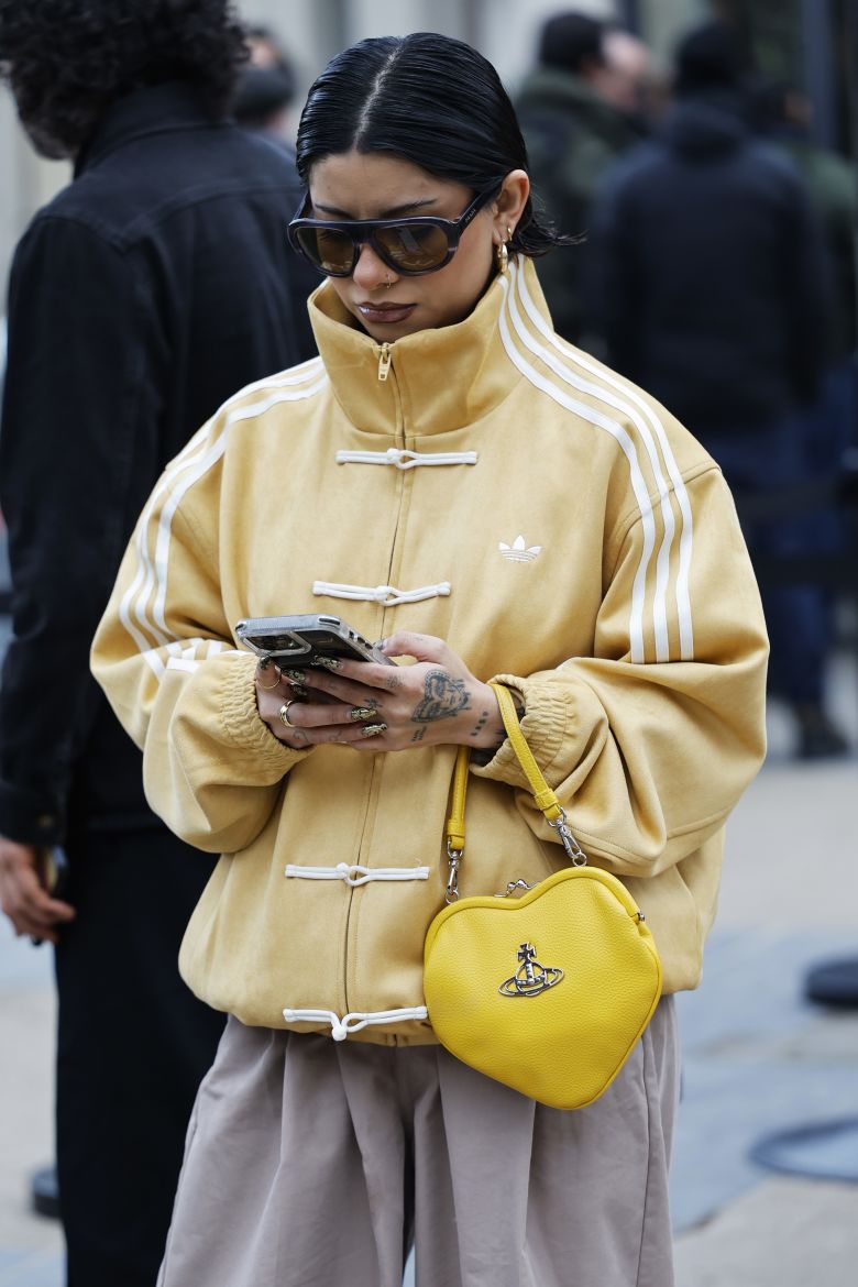 A guest wears dark sunglasses, a mustard yellow suede Adidas Tang jacket with white toggle closures, oversized beige cotton trousers, and a bright yellow Vivienne Westwood heart-shaped leather handbag, outside Jeanne Friot, during the Menswear Fall/Winter 2026-2027 as part of Paris Fashion Week on January 20, 2026 in Paris, France.