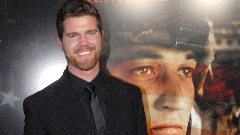 Actor Beau Wirick smiling on the red carpet at a movie premiere with a poster of Miles Teller in a soldier's helmet in the background