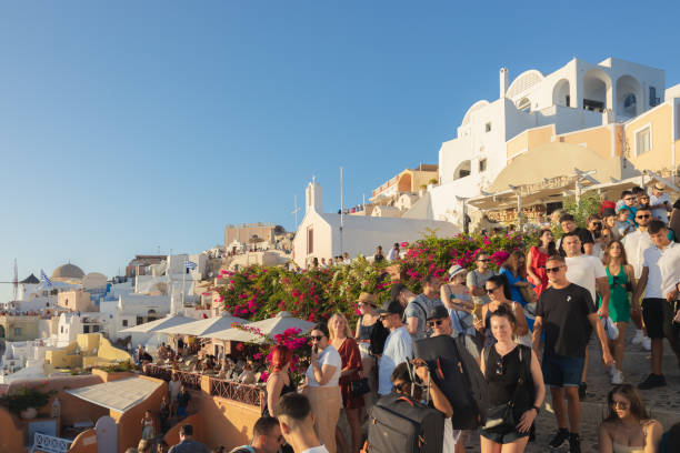 Crowds of tourists walking through Oia in Santorini with white buildings and bright flowers at sunset.
