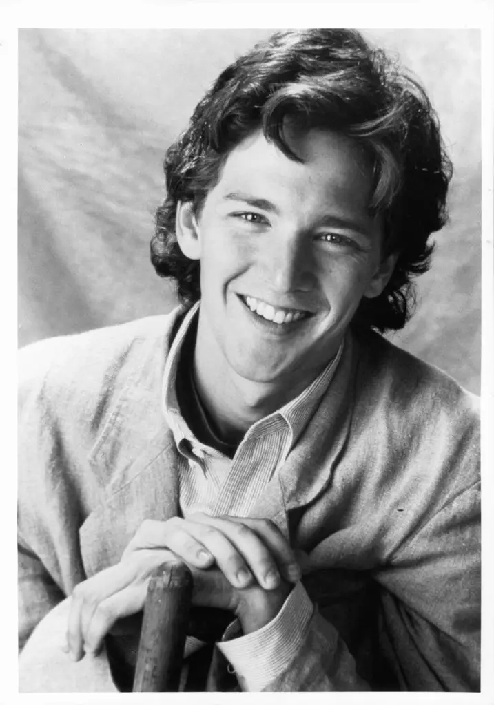 Andrew McCarthy leans against the back of a chair and holds his folded hands on its headrest.