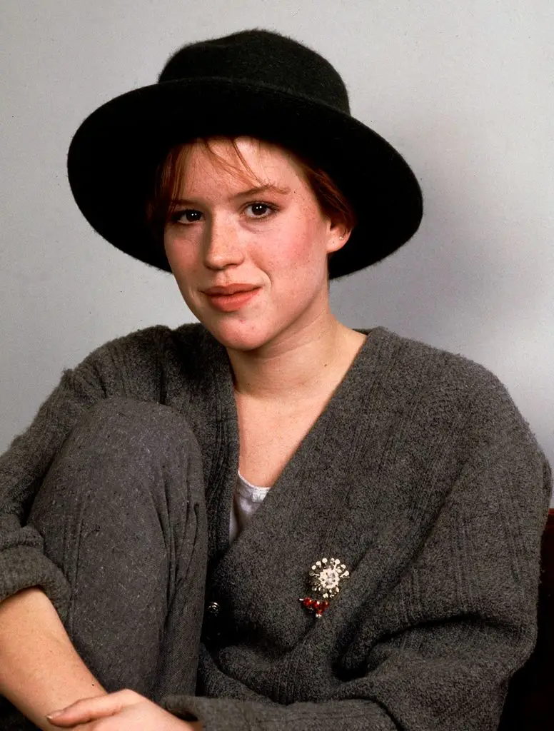 Portrait of Molly Ringwald in a dark grey cardigan and dark grey trousers, pulling her knee up to her chest with clasped hands, wearing a dark wide brim hat.