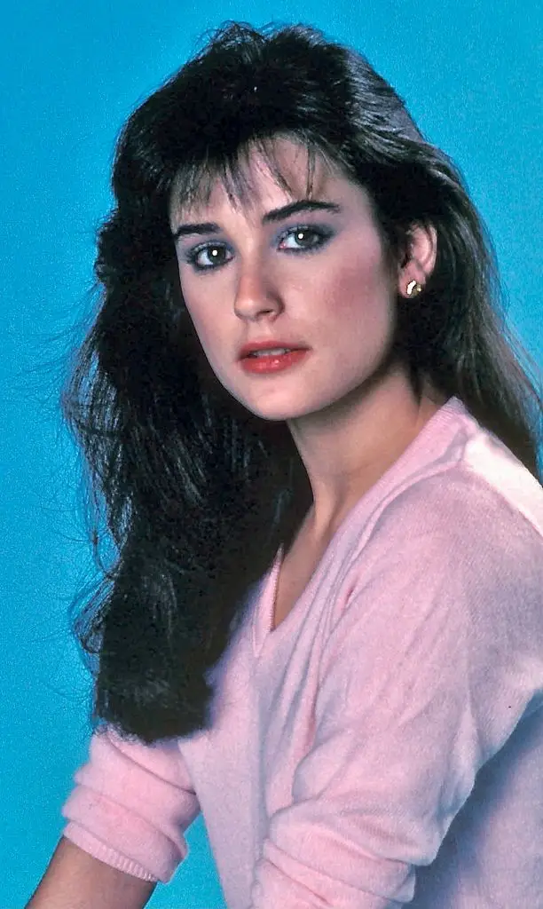 Demi Moore portrait. The actress faces her body to the left of the image and turns her head to her left to look at the camera. She wears a vneck pink jumper with the sleeves rolled to the elbow and poses in front of a blue background.