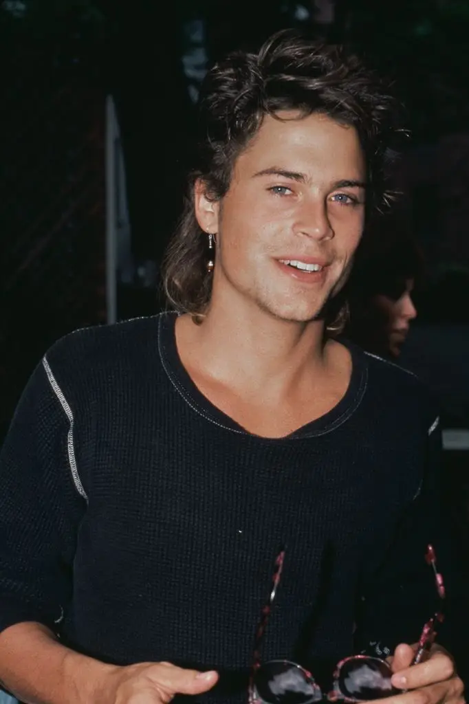American actor Rob Lowe wearing a dark crew neck top with white trim, and holding a pair of sunglasses with tortoiseshell frames.