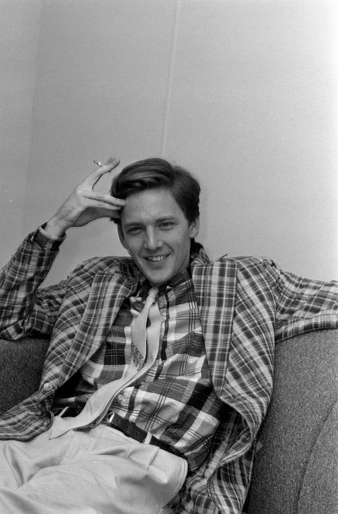 Andrew McCarthy sits back on a sofa, spreading his left arm along the back and bending his right arm at the elbow, pressing two fingers to his forehead and holding a cigarette. He wears a check jacket over a check shirt with light trousers.