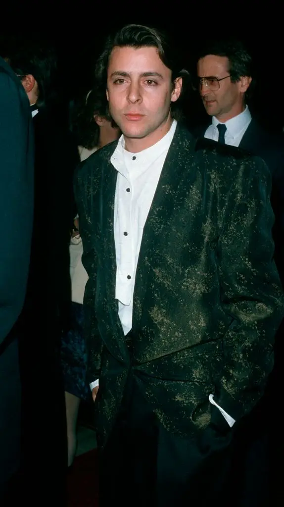 Judd Nelson in a green jacket over a white shirt with black trousers, holding his hands in his pockets and looking to the left of the image.