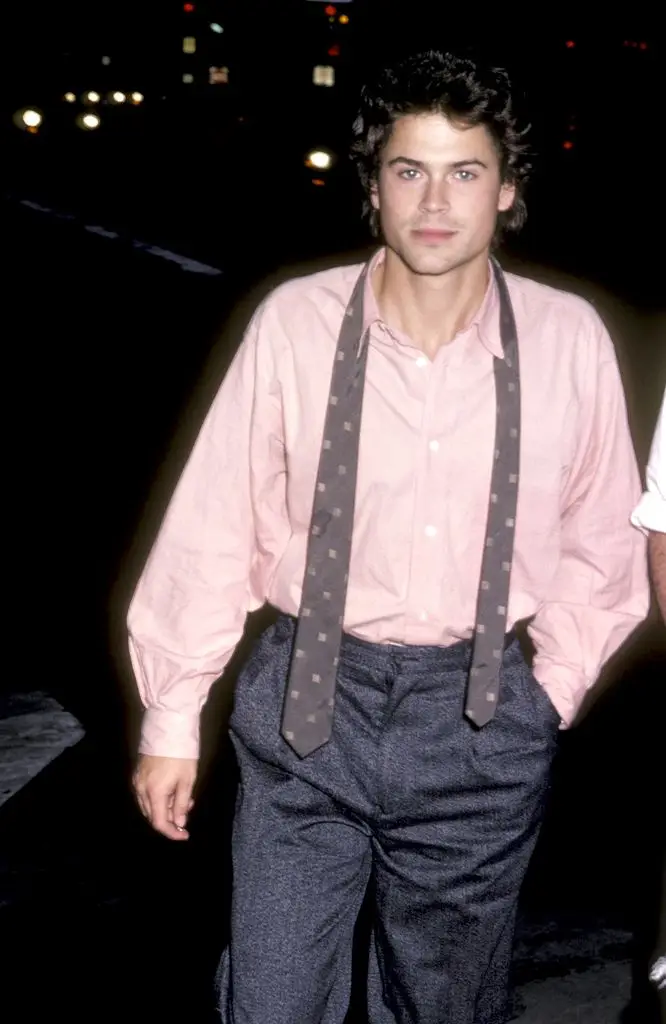 Rob Lowe wearing a pink shirt tucked into grey trousers with a dark spotted tie open around his neck. He walks forward and keeps his left hand in his pocket.