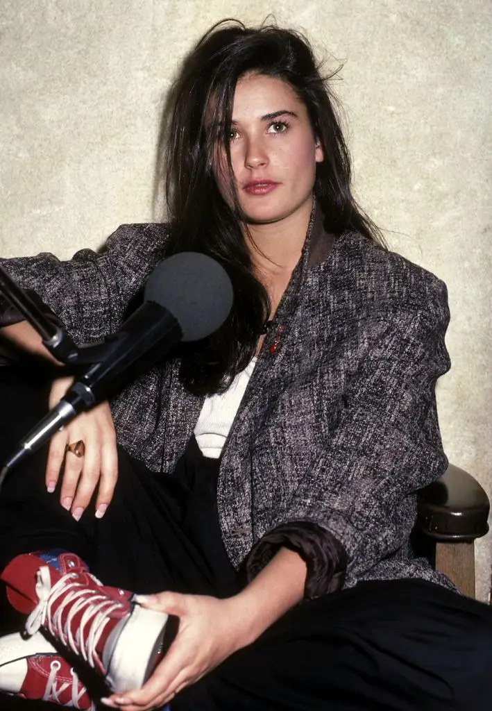 Demi Moore in a cream top, dark trousers, grey textured jacket and red converse, posing in front of a microphone with one hand resting on her right leg while the other hand holds her right shoe.