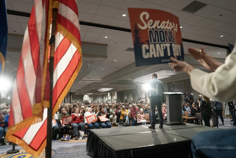 Graham Platner is seen from behind while standing on a temporary stage in front of a large audience.