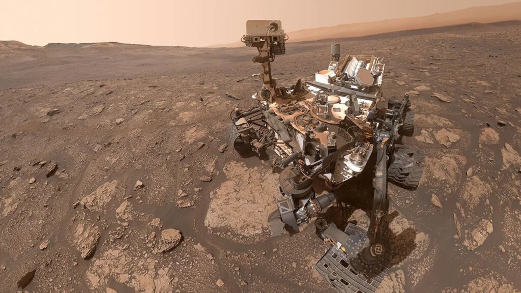 The six-wheeled Curiosity rover is positioned in the right side of the frame on a rugged, rock-strewn, brownish-red Martian plain. The rover’s articulated robotic arm and camera mast are raised. Distant hills and a hazy, dusty orange-brown sky are in the background.