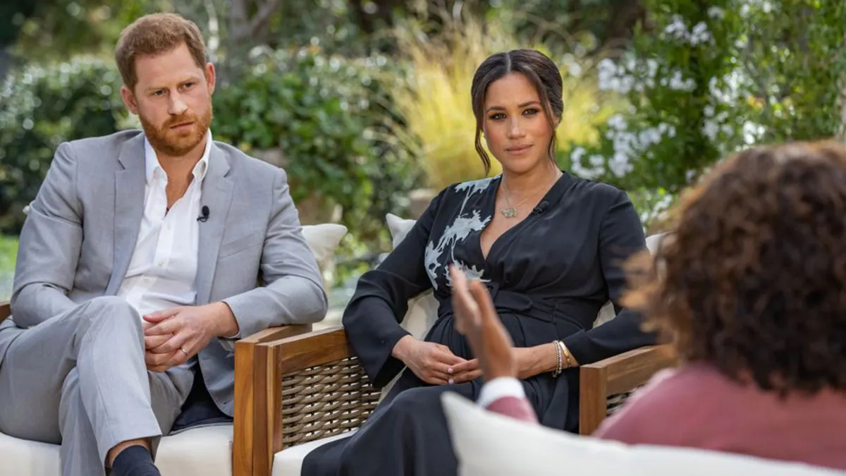 Prince Harry in grey suit and Meghan Markle in blue dress sitting across from Oprah Winfrey during interview