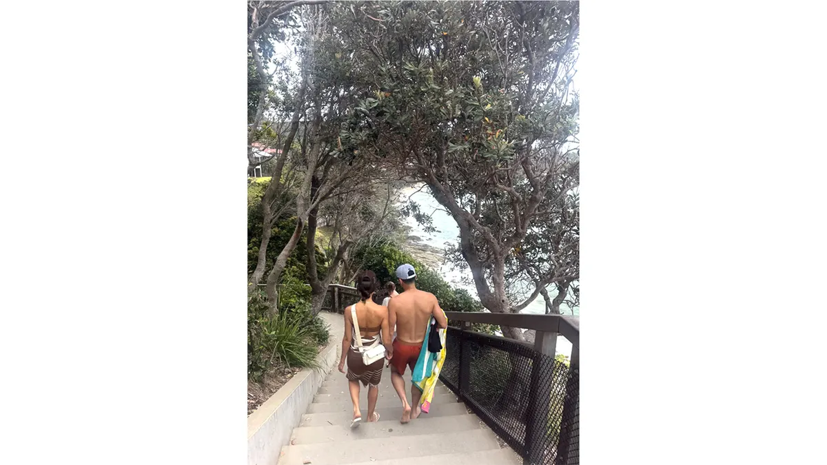 Jessica Alba and Danny Ramirez holding hands walking down a tree-lined staircase toward the beach.
