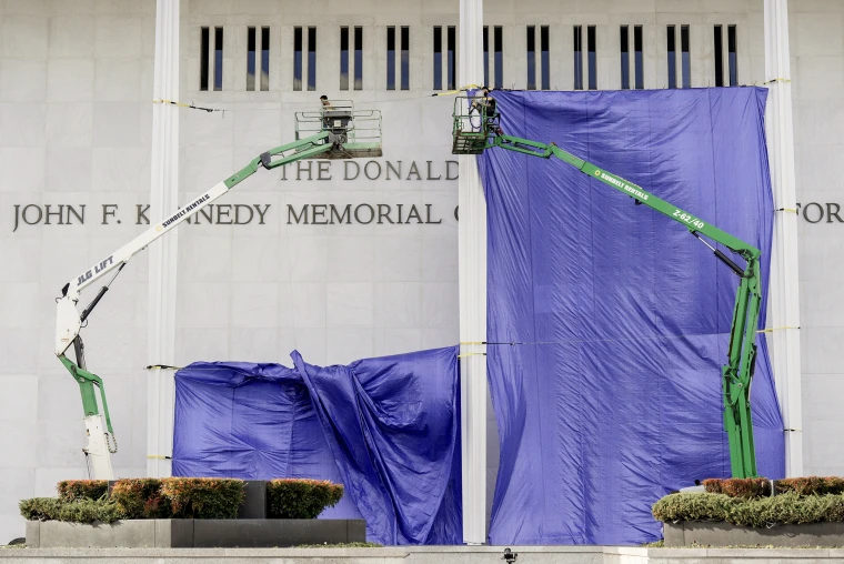 Workers in two hydraulic cranes are suspended above the ground in front of the Kennedy Center's facade, with a giant purple tarp stretched behind them.