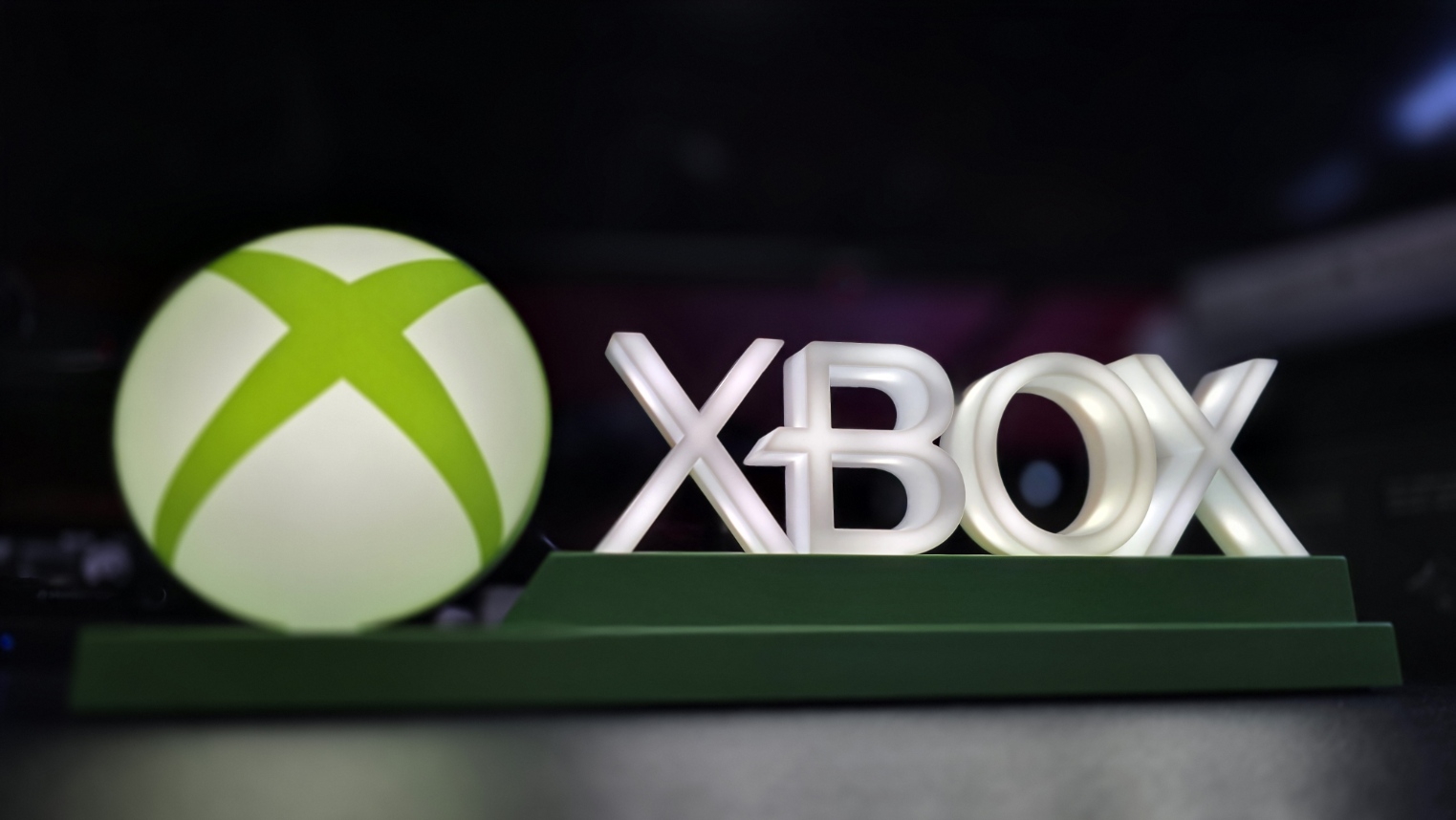 Illuminated Xbox logo display with glowing green sphere and white Xbox lettering against a dark blurred background.
