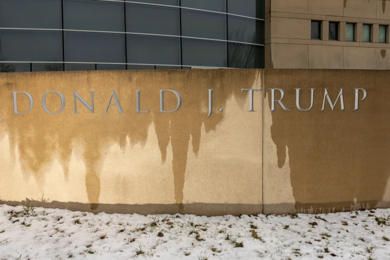 DONALD J. TRUMP etched into a wet stone facade, with snow covering the grass beneath it.