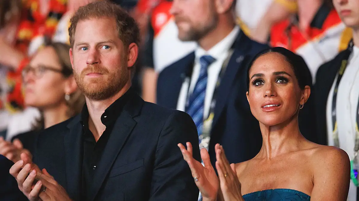 Prince Harry and Meghan Markle standing close to each other in an audience applauding and looking serious.