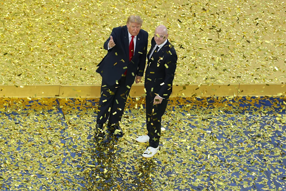 Two men stand amid gold confetti, one in a suit and tie, the other in a suit and sneakers, on a stage
