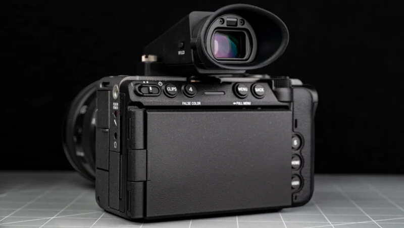 A digital camera with a closed flip-out screen is shown from the back, placed on a grid-patterned surface, with a black background.