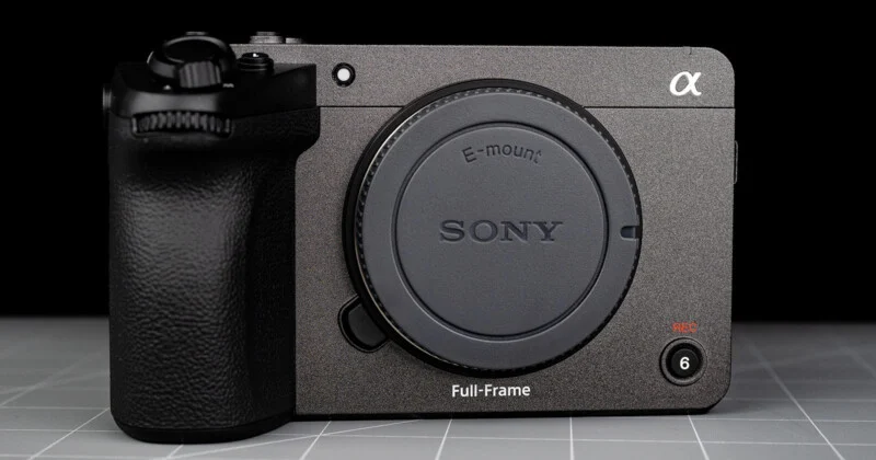 A close-up of a Sony Alpha full-frame mirrorless camera body with no lens attached, showing the E-mount and various control buttons on a gray grid-patterned surface.