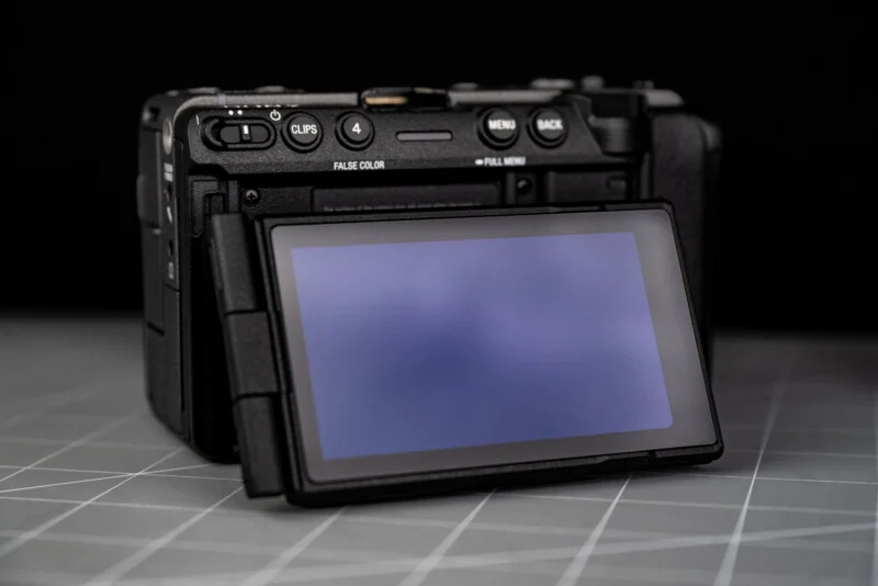 A digital camera with its rear LCD screen flipped out, facing forward. The camera sits on a gray surface with a grid pattern, and the background is black.