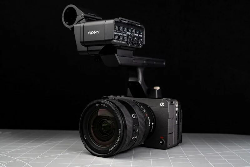 A professional Sony digital video camera with a large lens and external audio interface is placed on a gray grid-patterned surface against a black background.