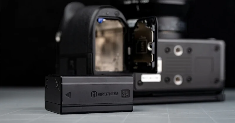 A camera with its battery compartment open is in the background, while a black InfoLITHIUM battery lies in the foreground on a flat surface.