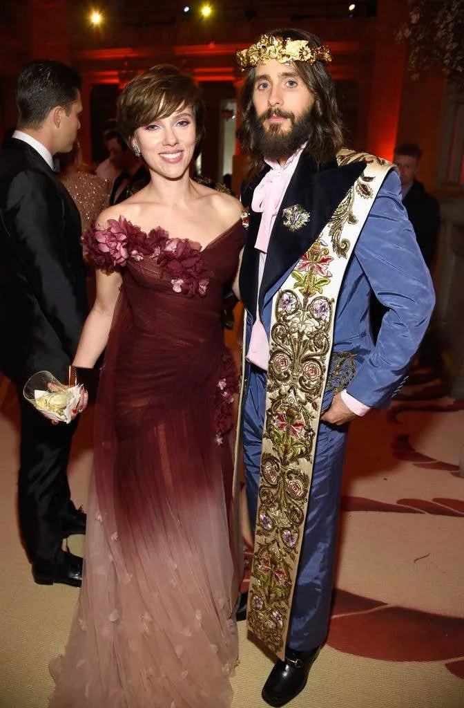 Scarlett Johansson in a dark red dress with her arm around Jared Leto, who's wearing a crown