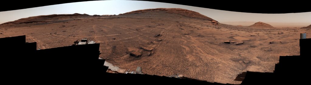A panoramic view of a sweeping Martian plain covered in patterned fractures leading toward distant hills showcases the terrain explored by NASA's Curiosity rover to investigate the planet's past environment.