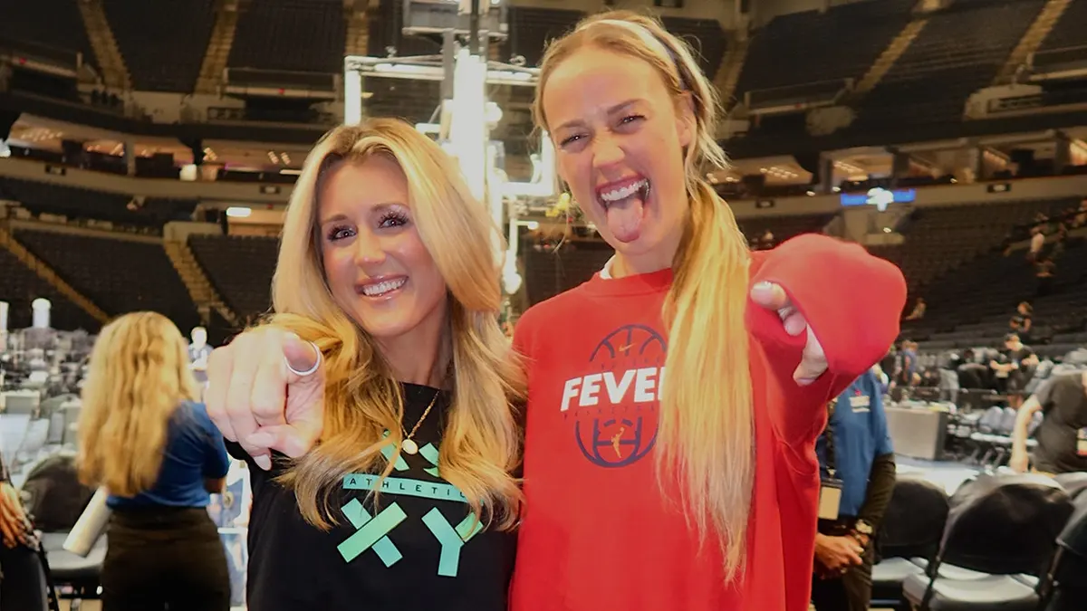 Riley Gaines and Indiana Fever guard Sophie Cunningham posing together on the basketball court, smiling and pointing at the camera inside a packed arena.