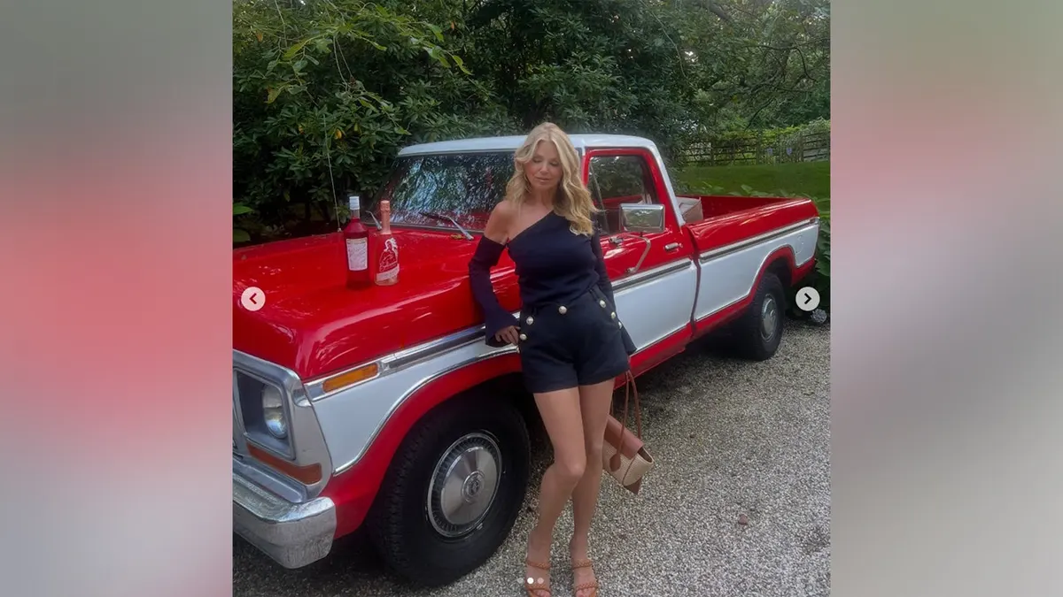 Christie Brinkley looking down as she leans against a car.