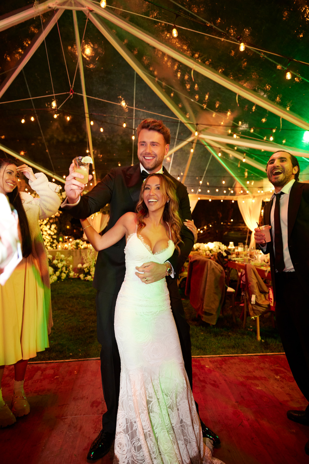 Bride and groom smiling and dancing at night under a clear tent decorated with string lights, surrounded by happy guests at an outdoor wedding reception.