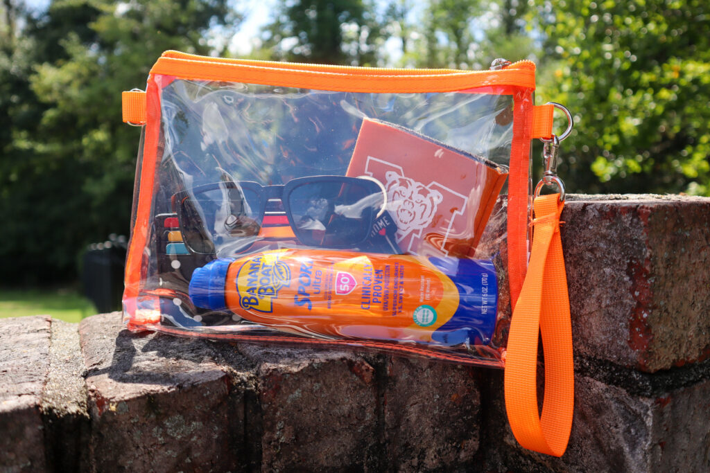 Clear orange-trimmed bag on brick holds sunglasses, sunscreen, a small notebook, and striped cardholder outdoors.