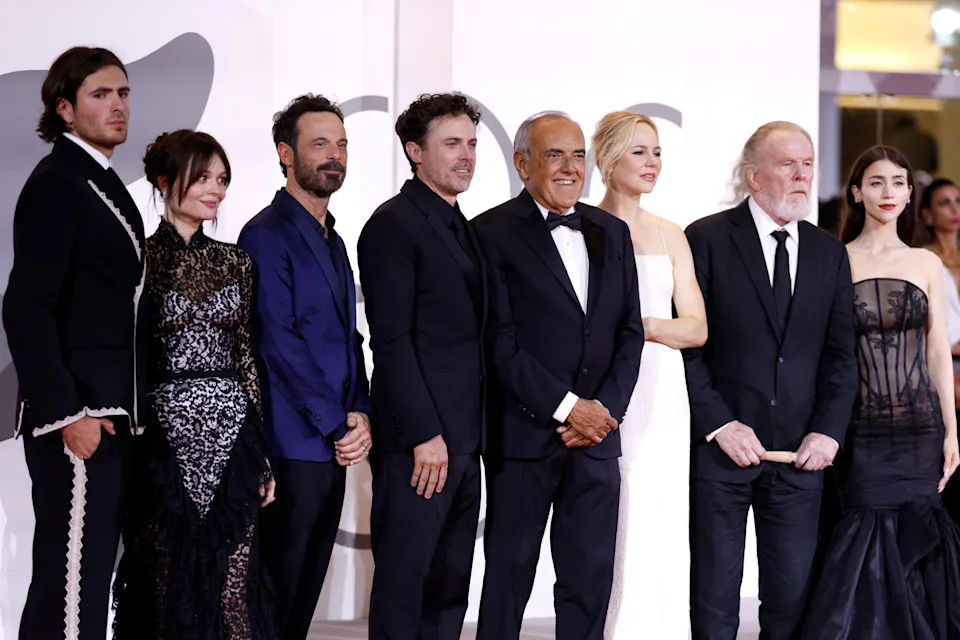 Group of elegantly dressed people posing, including several in formal attire with one in a tuxedo and a gown, at an event