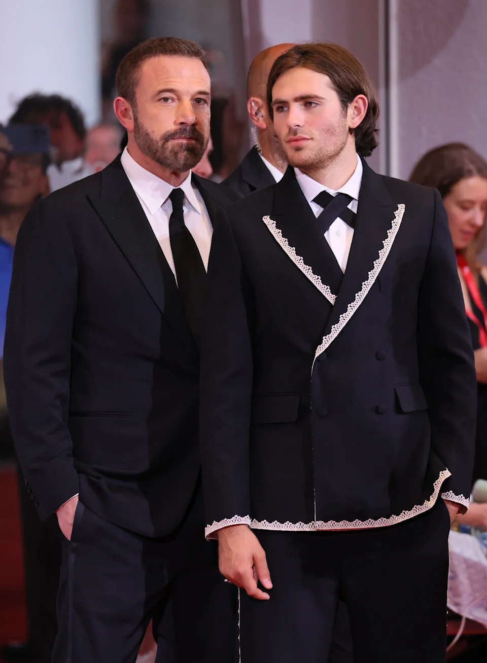 Two individuals in stylish suits on a red carpet at an event, engaging with onlookers, showcasing sleek formal attire and confident expressions