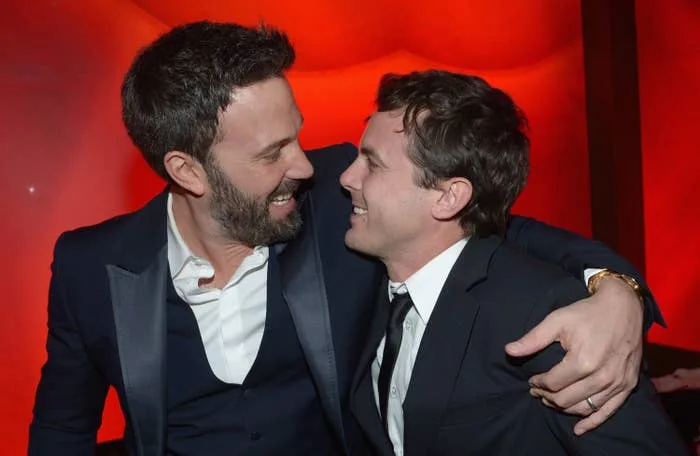 Two people in suits smile warmly at each other, embracing in a friendly manner against a vibrant background
