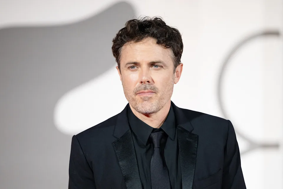 A person in a sleek suit poses on a red carpet, with a blurred background