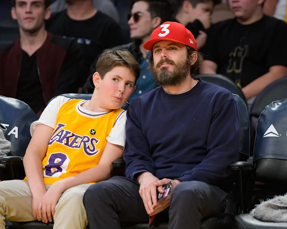 A person and a child sit courtside at a basketball game. The child wears a Lakers jersey, and the person wears a cap with the number 3 on it