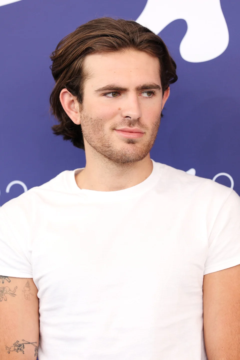 Person with medium-length hair, wearing a plain white t-shirt, seated against a blue backdrop at an event