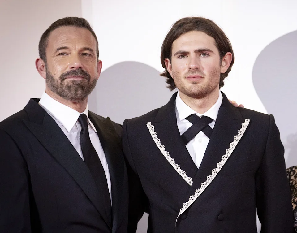 Three people on a red carpet; two men in suits, one with lace trim, and a woman in a lace dress