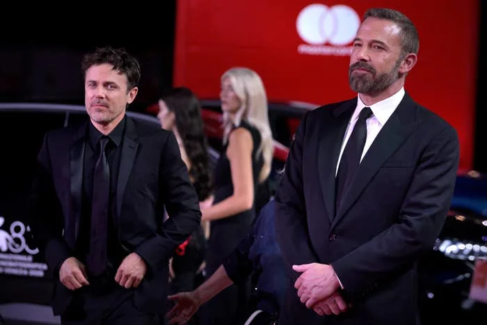 Two men in suits on a red carpet, standing in front of a backdrop with logos, surrounded by event attendees