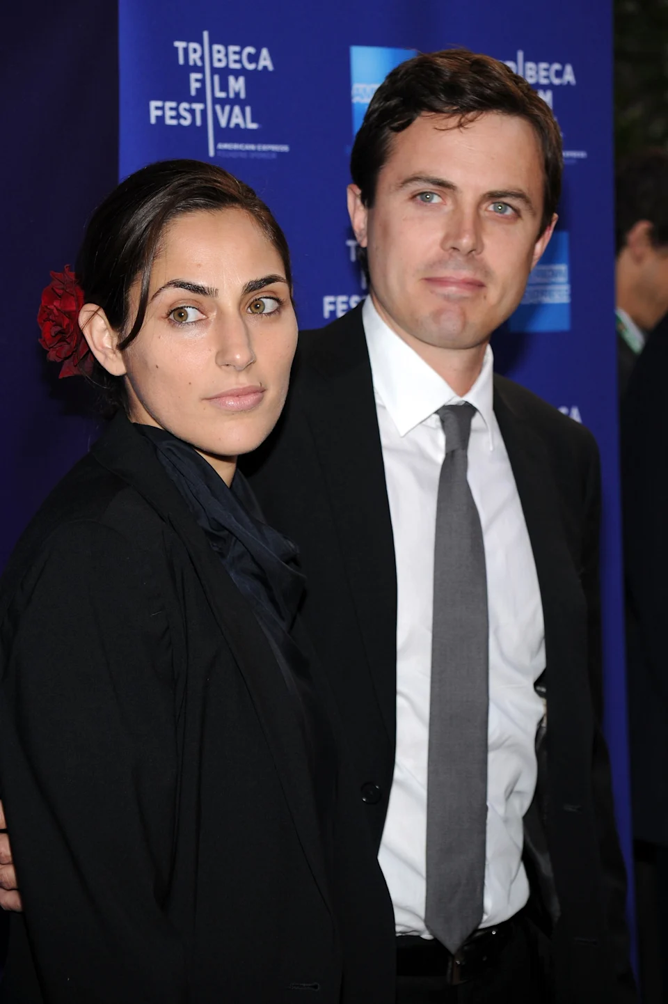 Two people posing in formal attire at the Tribeca Film Festival, one wearing a suit and tie, and the other in a blazer with a floral hair accessory